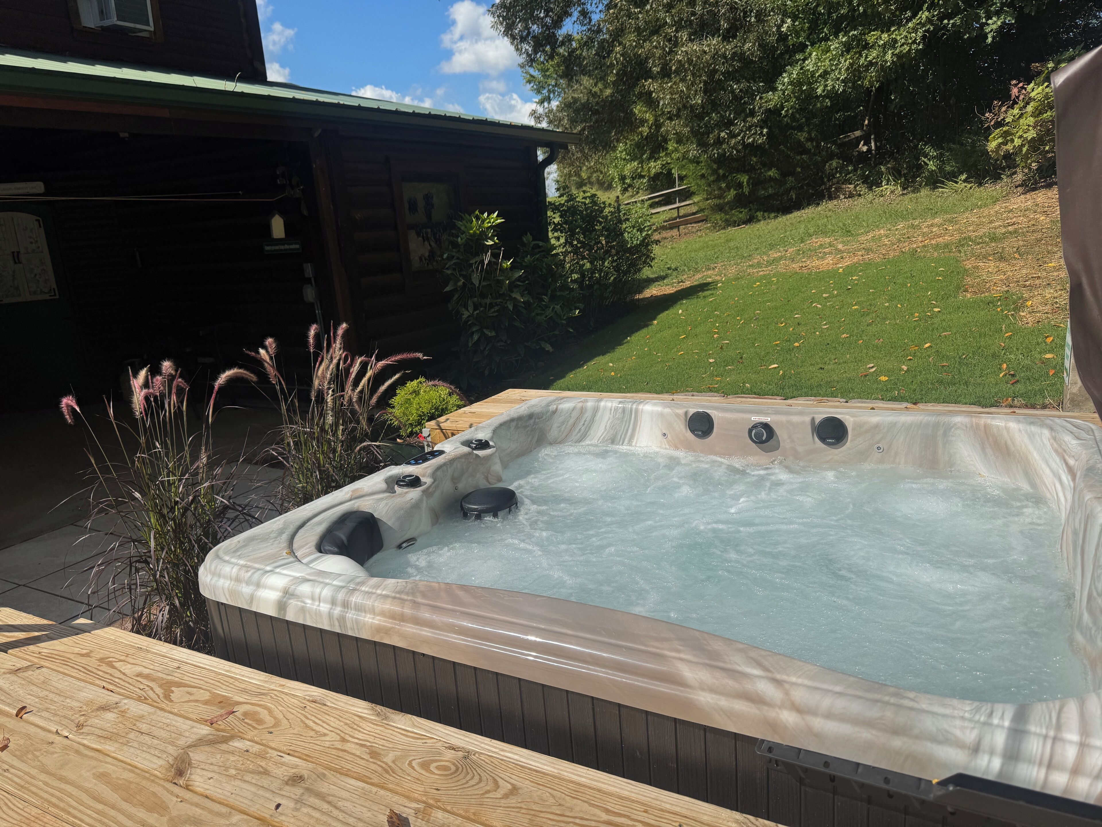 Un jacuzzi dans un jardin, avec une terrasse en bois, entouré de verdure et avec un bâtiment visible au loin.