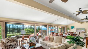 A spacious living room with a large window, a sofa, a coffee table, and a dining area with chairs and a table. - Spacious 2BR Best Ocean Views -Kapalua Golf Villas 11P 5-6 (Lahaina)