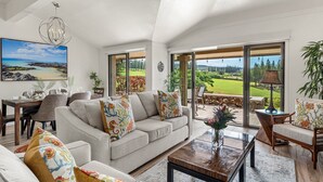 Ein geräumiges Wohnzimmer mit einer großen Couch, einem Couchtisch und einem Essbereich mit Stühlen und einem Tisch. Ein großes Fenster bietet Blick auf einen Golfplatz.