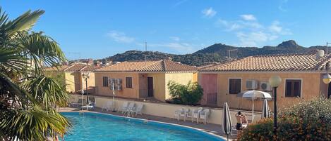 A swimming pool with lounge chairs, a palm tree, and buildings with tiled roofs in the background.