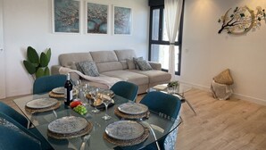Ein modernes Wohnzimmer mit einem Glastisch für sechs Personen, einem beigefarbenen Sofa mit gemusterten Kissen und einem großen Fenster mit weißen Vorhängen.