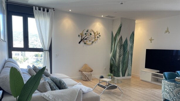Ein modernes Wohnzimmer mit einer Couch, einem Glastisch, einem Fernseher und einem großen Fenster mit Vorhängen.