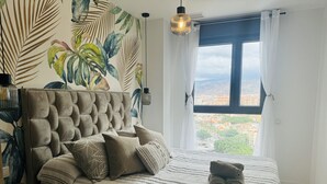 Ein Schlafzimmer mit einem gepolsterten Kopfteil, gestreifter Bettwäsche und Blick auf eine Stadtlandschaft durch ein Fenster.