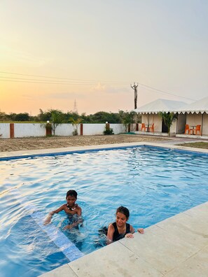 A swimming pool with two children playing, surrounded by a white wall and a building with outdoor seating.