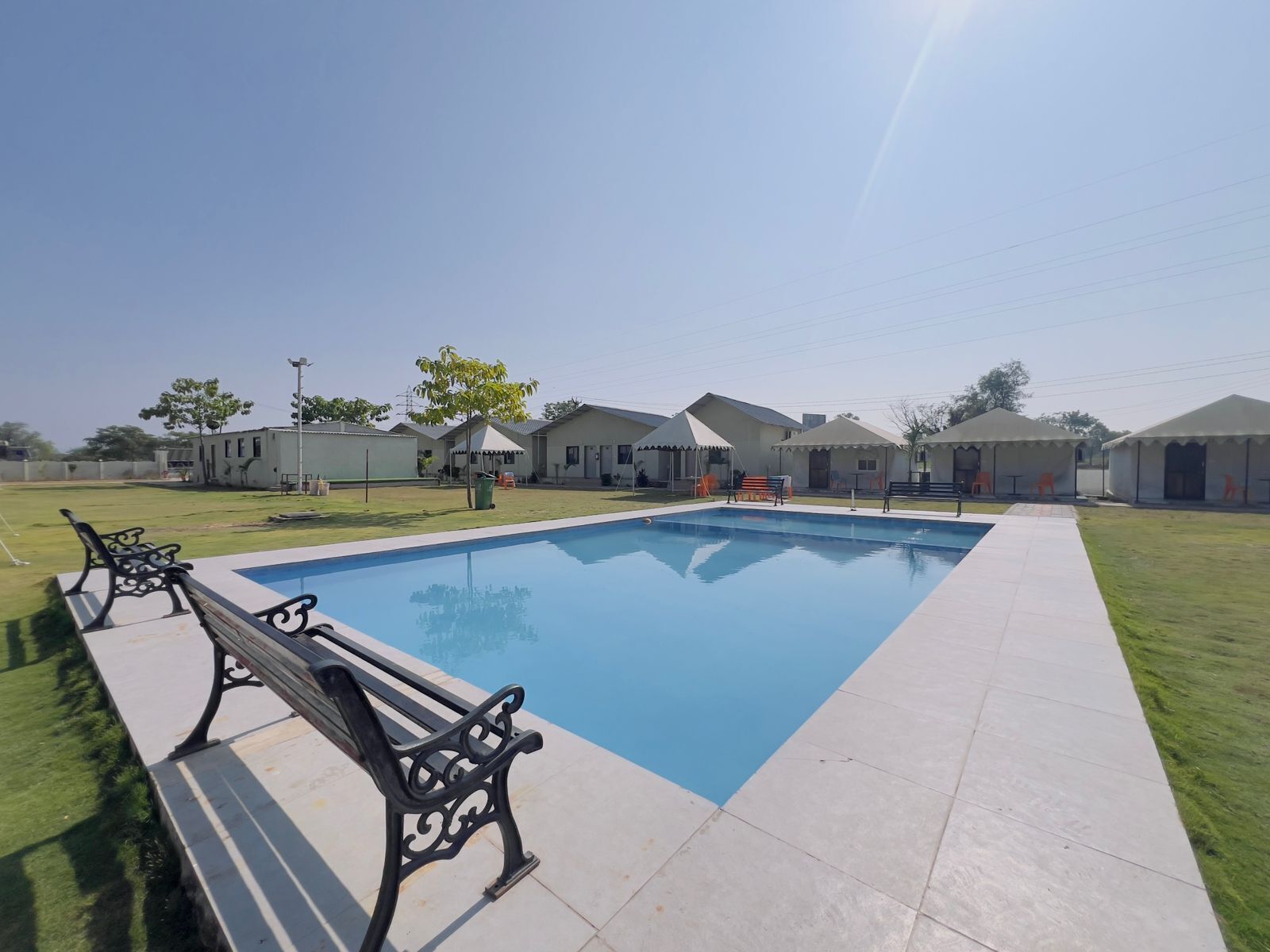 Una piscina con plataforma de baldosas, rodeada de estructuras cubiertas y un área verde.