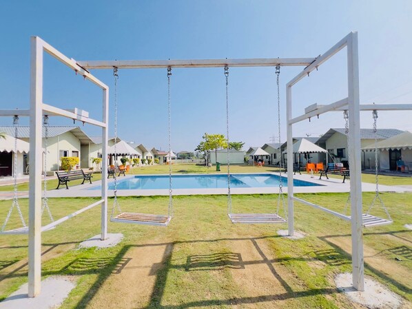 A pool area with swings, a clear sky, and a row of houses in the background. - Unity Sunrise Resort Statue of Unity Ekta Nagar (Garudeshwar)