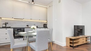 A modern kitchen with white cabinets, a round glass table, grey upholstered chairs, and a wooden TV stand.