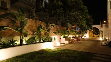 A well-lit outdoor area with a dining table and chairs, surrounded by lush greenery and a white planter.