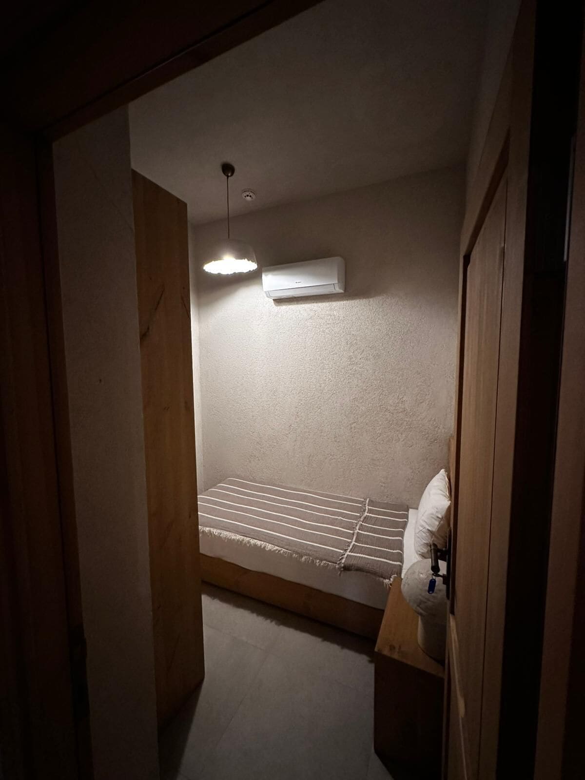 A small, dimly lit room with a single bed, a wall-mounted air conditioner, and a hanging light fixture.