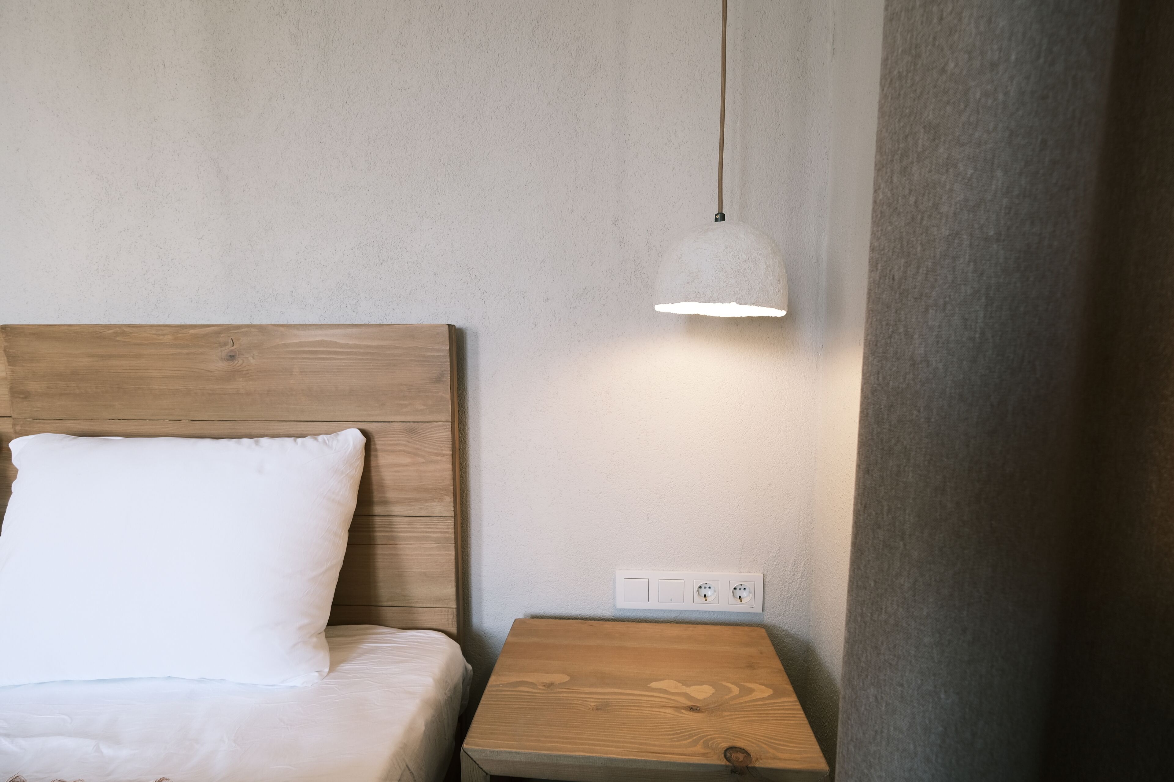 Una cama bien hecha con cabecera de madera y almohada blanca, una mesita de noche de madera con lámpara, y un enchufe en la pared.