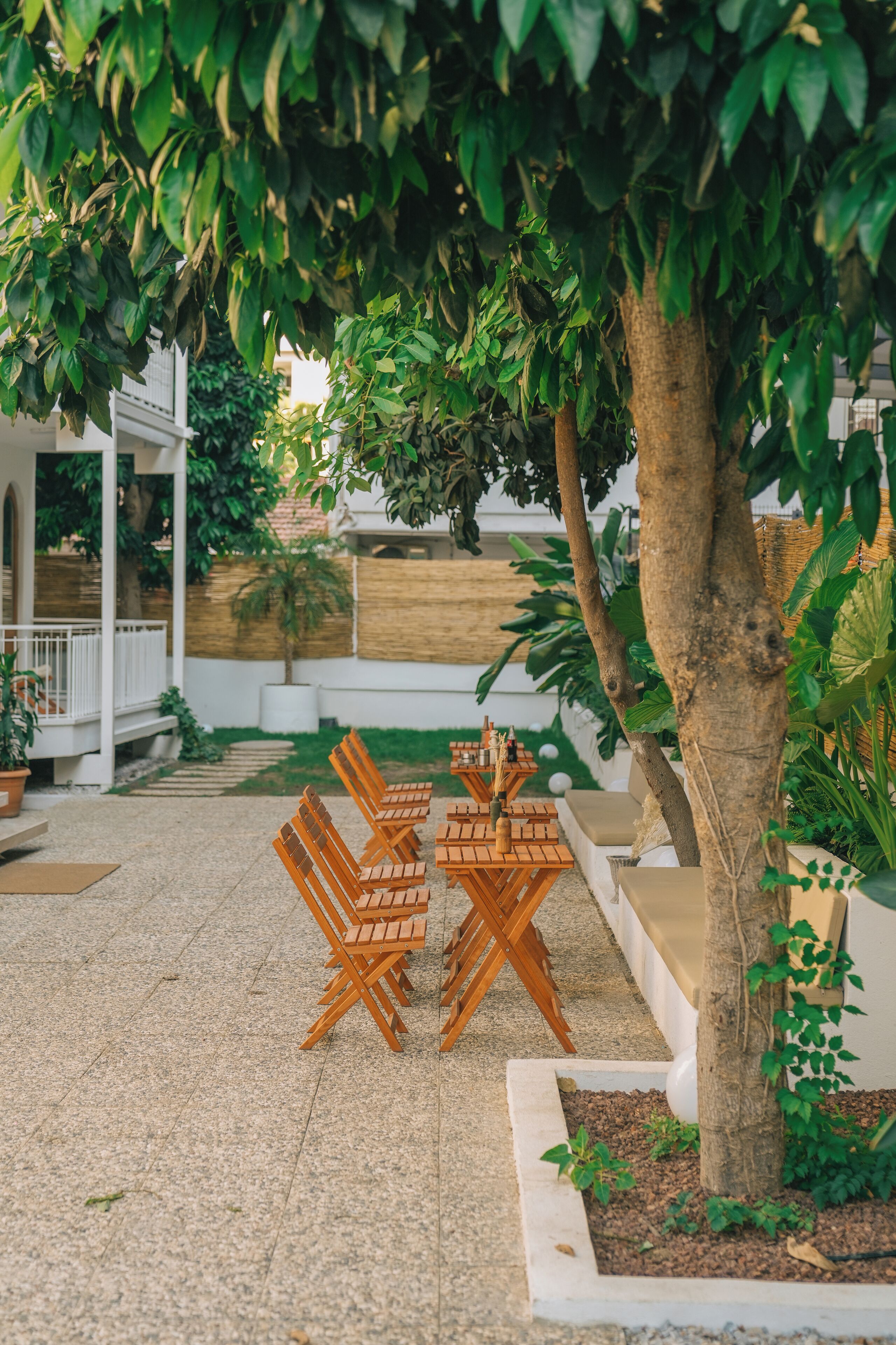 Un espace extérieur aménagé avec des chaises et des tables en bois, entouré de verdure et avec un bâtiment blanc en arrière-plan.