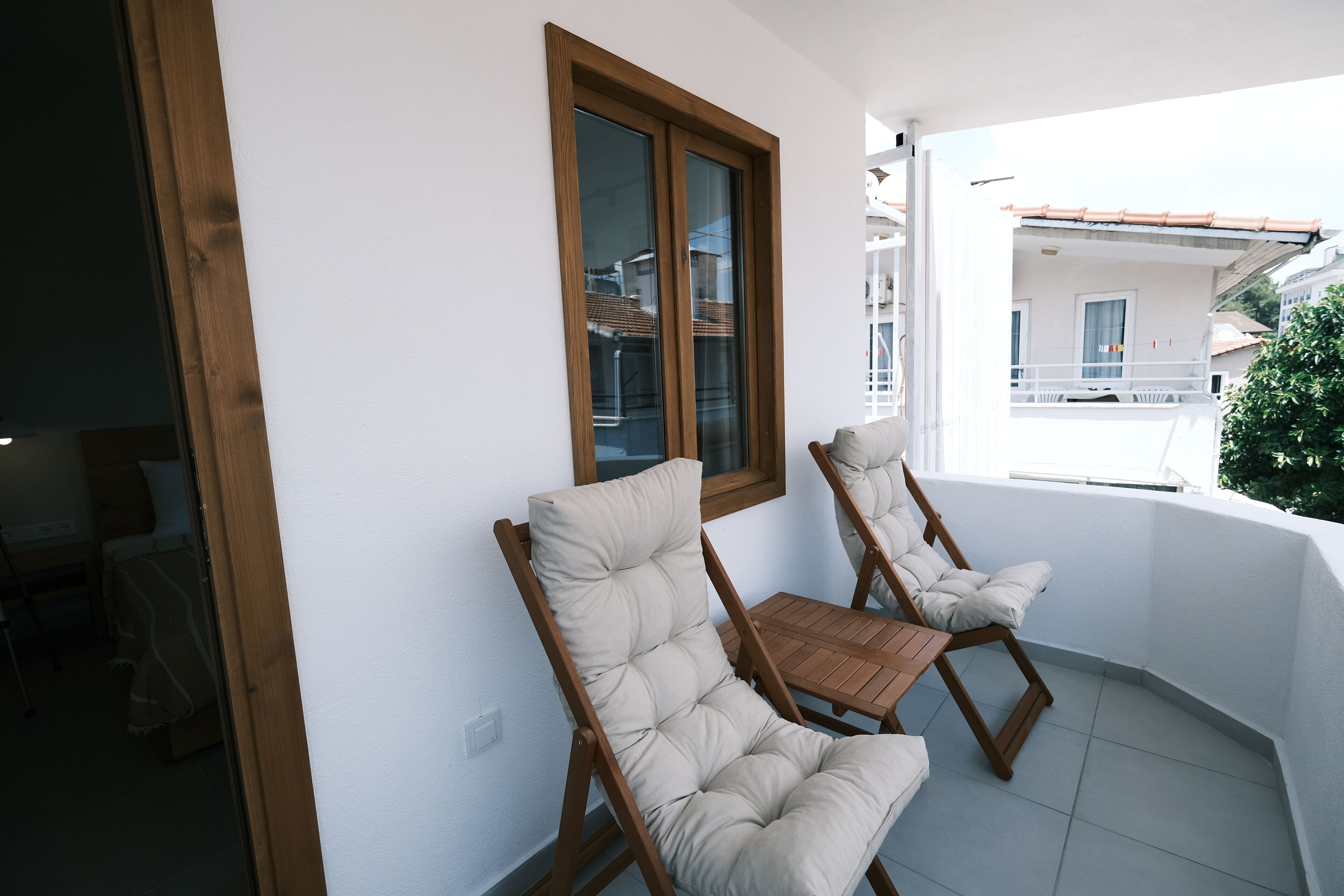 Un balcón con dos sillas acolchadas y una mesa de madera, una ventana con marco de madera, y vista a un edificio de la calle.