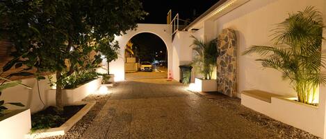 Vista nocturna de un sendero pavimentado que conduce a la entrada de un edificio, flanqueado por macetas y un muro de piedra.