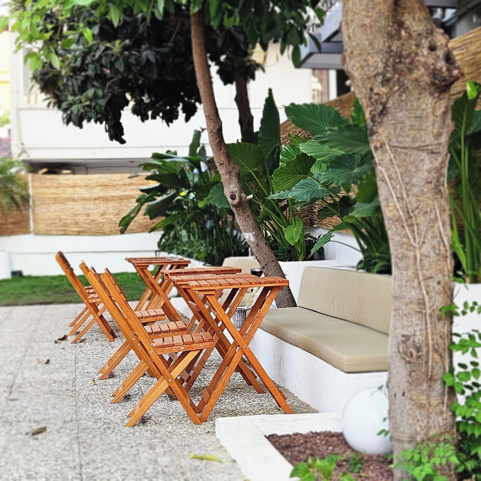 A patio area with wooden chairs and a sofa, surrounded by greenery and a white wall.