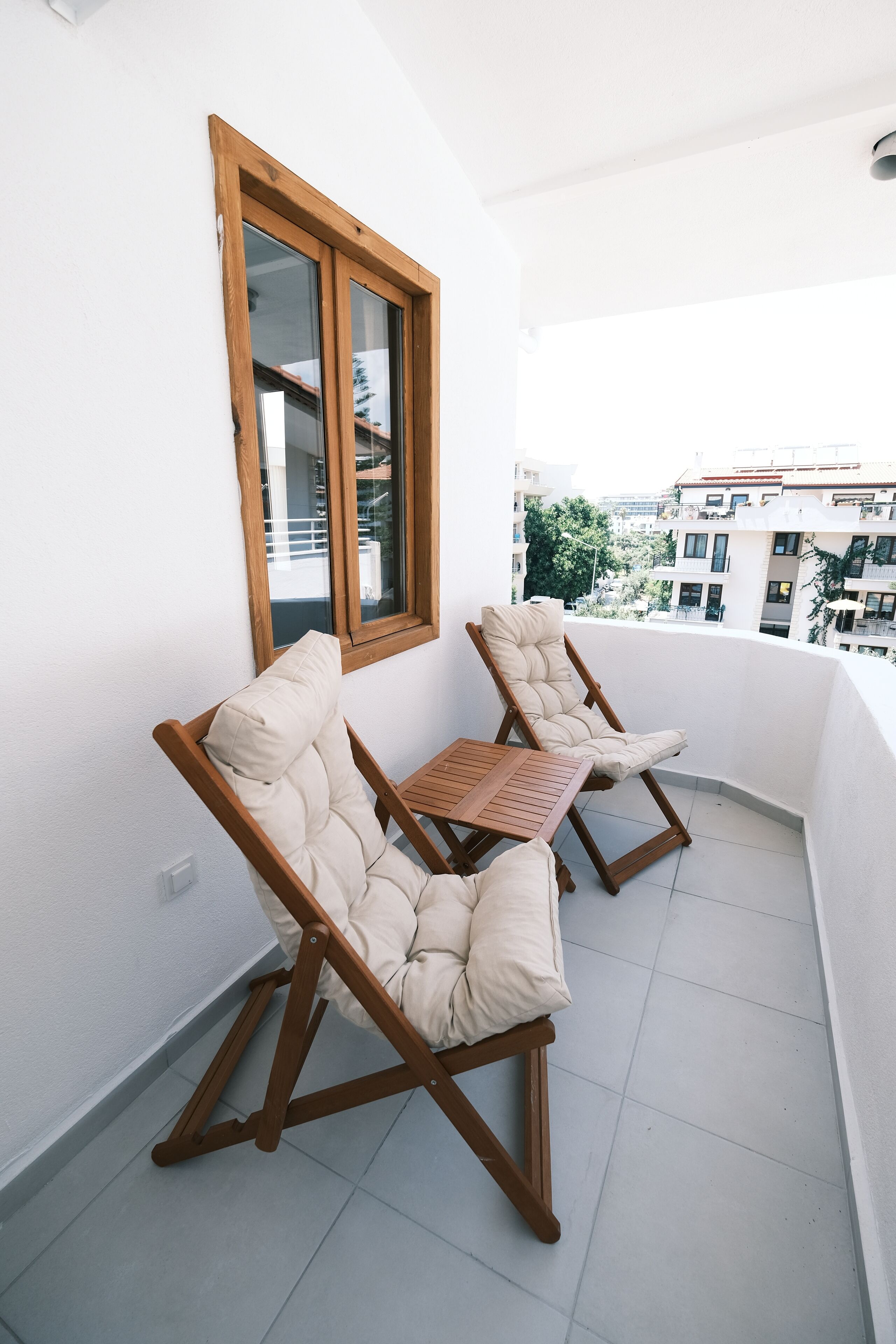 A balcony with two cushioned chairs and a small table, white walls, and a view of buildings.