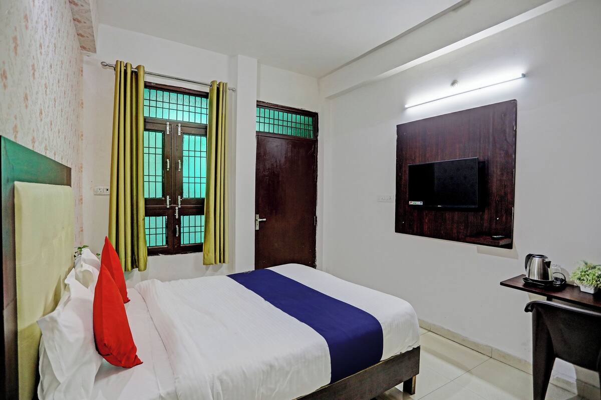 A hotel room with a bed, a TV mounted on the wall, a desk with a kettle, and curtains on the window.
