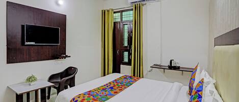 Habitación de hotel con cama, escritorio, silla, televisor montado en la pared, aire acondicionado y ventana con cortinas.