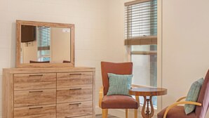 Une pièce comprenant une commode en bois, deux chaises, une petite table ronde et une fenêtre avec des stores.