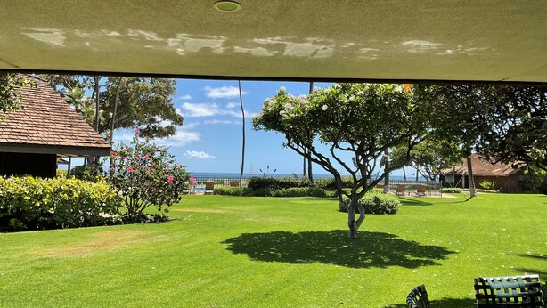A view from inside a building looking out to a well-maintained lawn with a tree, a small table, and a chair. - Ma'alaea Surf Unit F3-1 BR Ground Floor Beachfront Condo, Wi-Fi, AC, Pool, TV (Kihei)