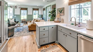 A modern kitchen with grey cabinets, a white countertop, and a stainless steel dishwasher. There is a dining area with a sofa and a coffee table. - Just Peachy (Carlton landing)