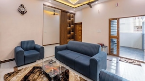 Uma sala de estar moderna com um sofá azul, uma poltrona combinando e uma mesa de centro de vidro. Uma porta de madeira e um ventilador de teto são visíveis.