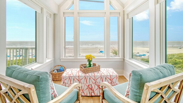 Ein sonnendurchflutetes Zimmer mit großen Fenstern mit Blick auf einen Strand, mit einem Korbwäschesessel, einem gemusterten Hocker und zwei Holzstühlen.