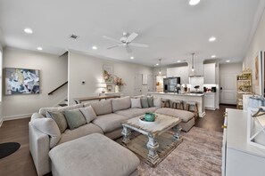 A modern living room with a sectional sofa, a coffee table, and a dining area with a kitchen in the background. - Coastal Tide - Luxury Townhome - Book for Football Season! (Tuscaloosa)