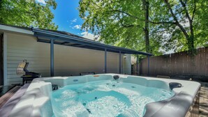Ein Whirlpool in einem Garten mit überdachter Terrasse, Holzzaun und Bäumen im Hintergrund.