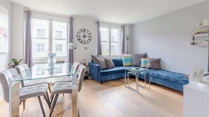 A modern living room with a glass dining table, a blue sofa, and a large wall clock. - Chic 1BD Flat in the Heart of Islington! (Greater London)