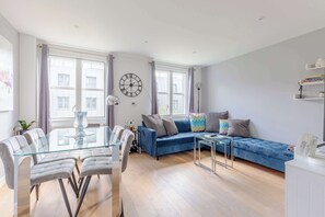 A modern living room with a glass dining table, a blue sofa, and a large wall clock. - Chic 1BD Flat in the Heart of Islington! (Greater London)