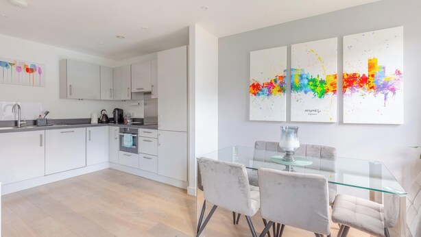 A modern kitchen with white cabinets, a glass dining table, and a cityscape artwork on the wall. - Chic 1BD Flat in the Heart of Islington! (Greater London)