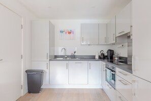 A modern kitchen with white cabinetry, a stainless steel sink, and a black trash bin. - Chic 1BD Flat in the Heart of Islington! (Greater London)