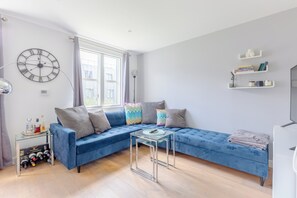 A modern living room with a blue sectional sofa, a glass coffee table, a wall clock, and a window with curtains. - Chic 1BD Flat in the Heart of Islington! (Greater London)