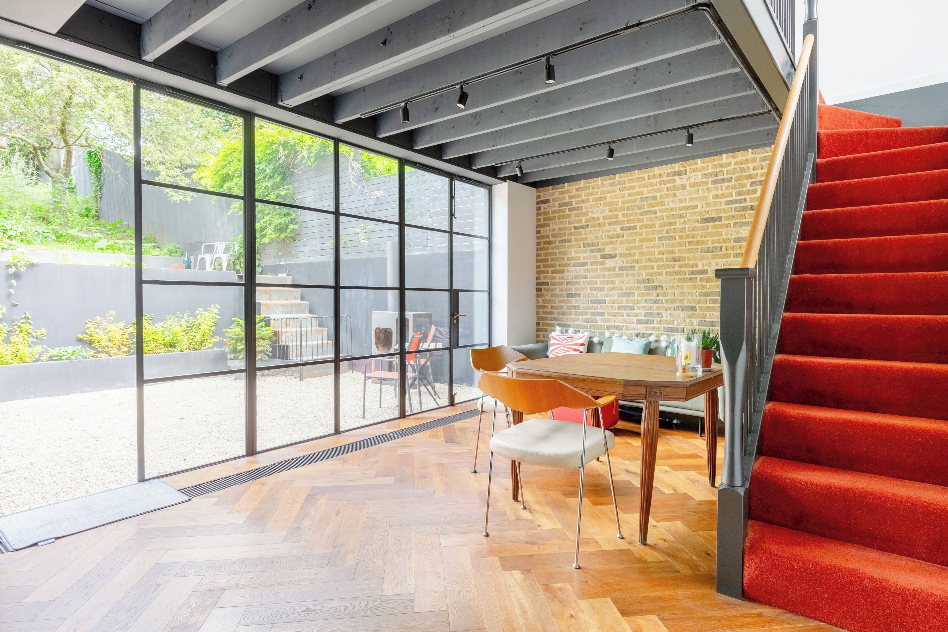 A modern interior with a herringbone wooden floor, a brick wall, and a red carpeted staircase.