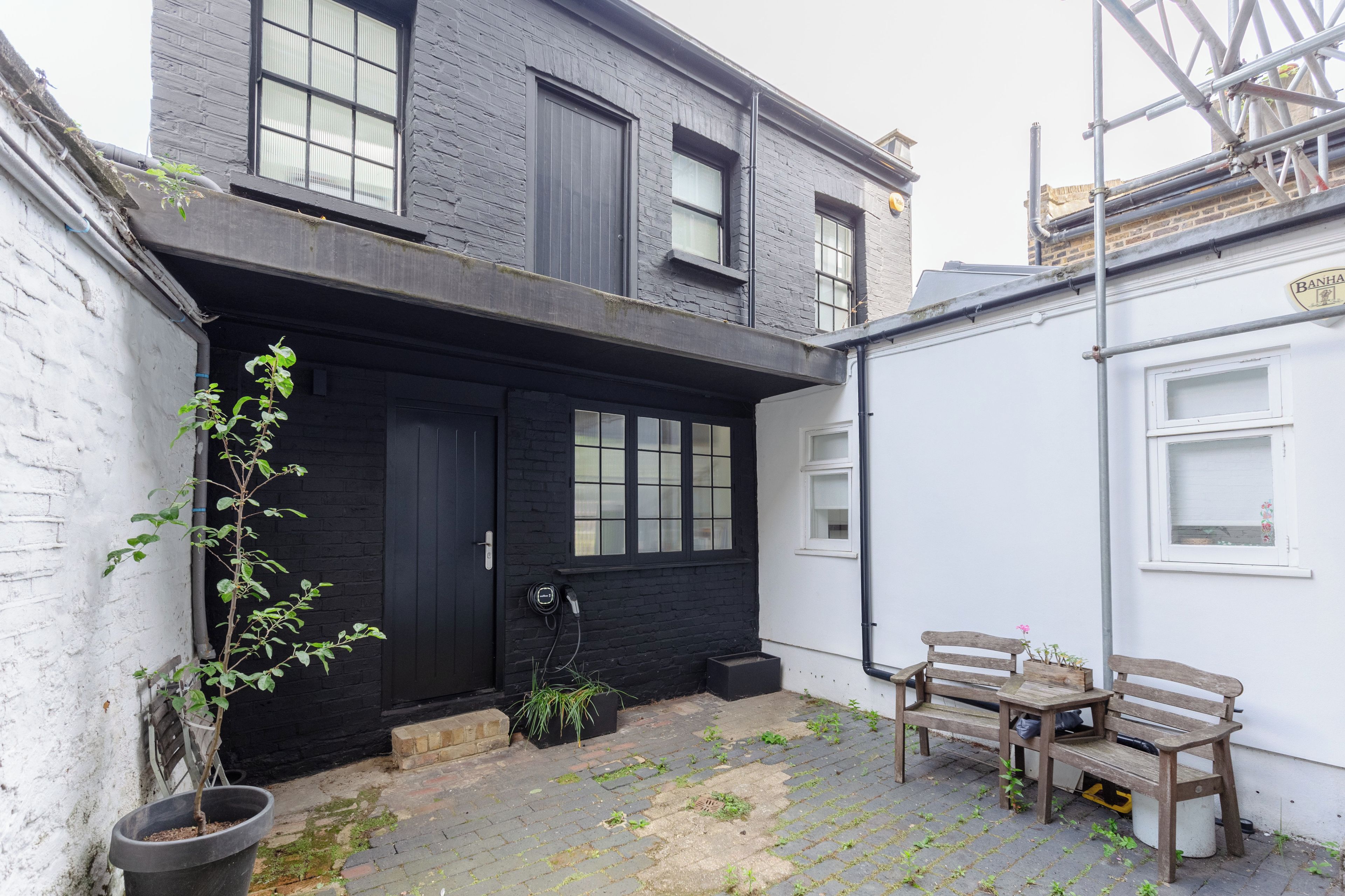 Un bâtiment à deux étages avec une porte d’entrée noire, un petit jardin agrémenté d’un banc et de plantes en pot, et un mur extérieur blanc.
