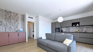 Un salon moderne avec un canapé gris, un meuble rose et un coin cuisine doté de meubles gris et d’un crédence à motifs.