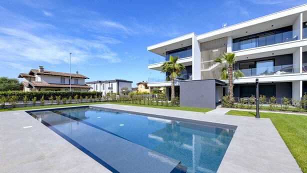 Un espace piscine moderne doté d’une piscine rectangulaire, situé à côté d’un bâtiment contemporain à deux étages, agrémenté de balcons et d’une pelouse bien entretenue.