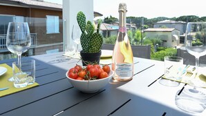 Une table avec un saladier de tomates, une bouteille de champagne et des verres à vin vides, le tout installé sur un balcon d’où l’on peut admirer des maisons et la verdure.