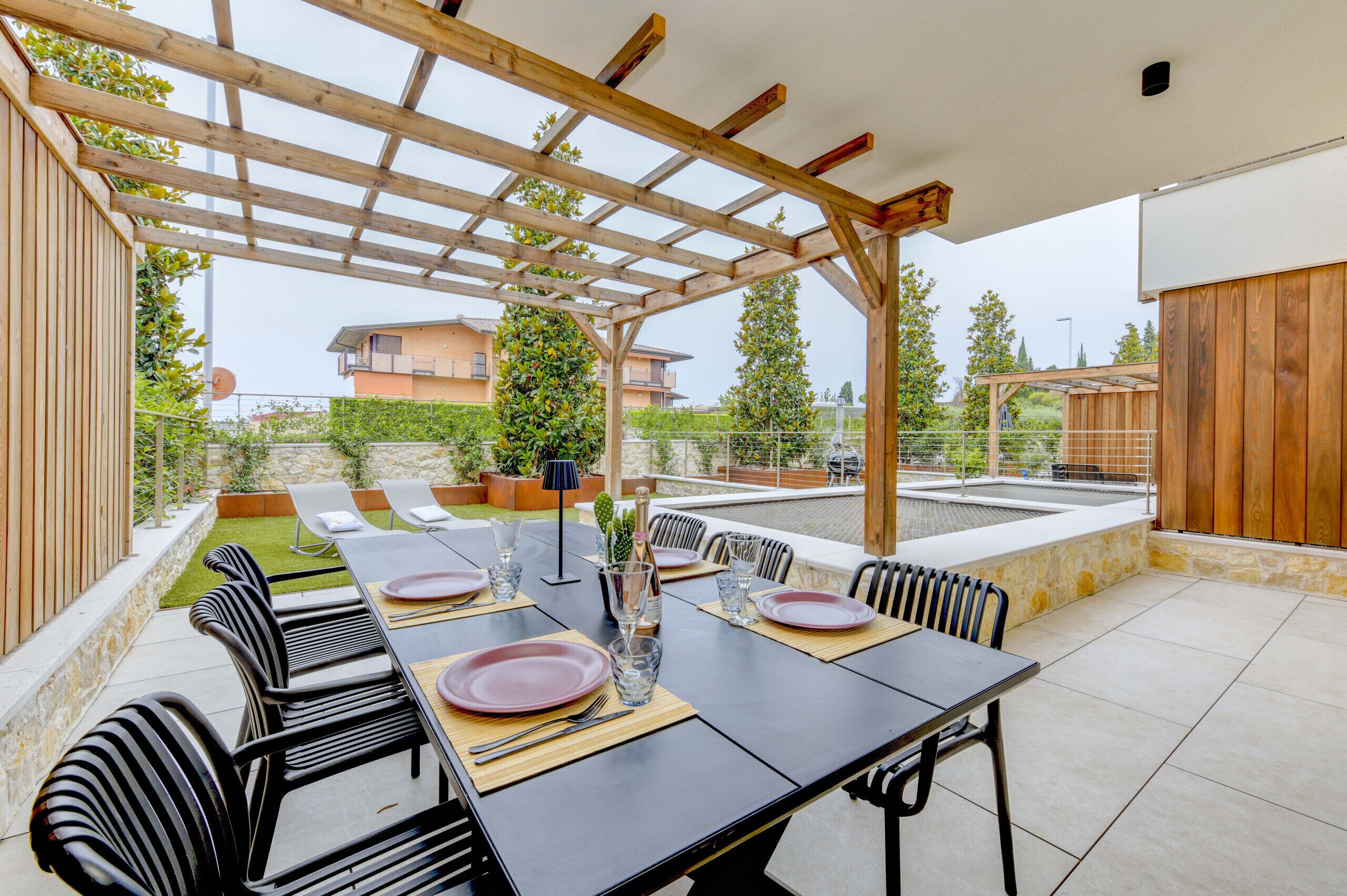 Een terras buiten met een houten pergola, een eettafel gedekt voor zes personen en een stenen muur begroeid met groen.