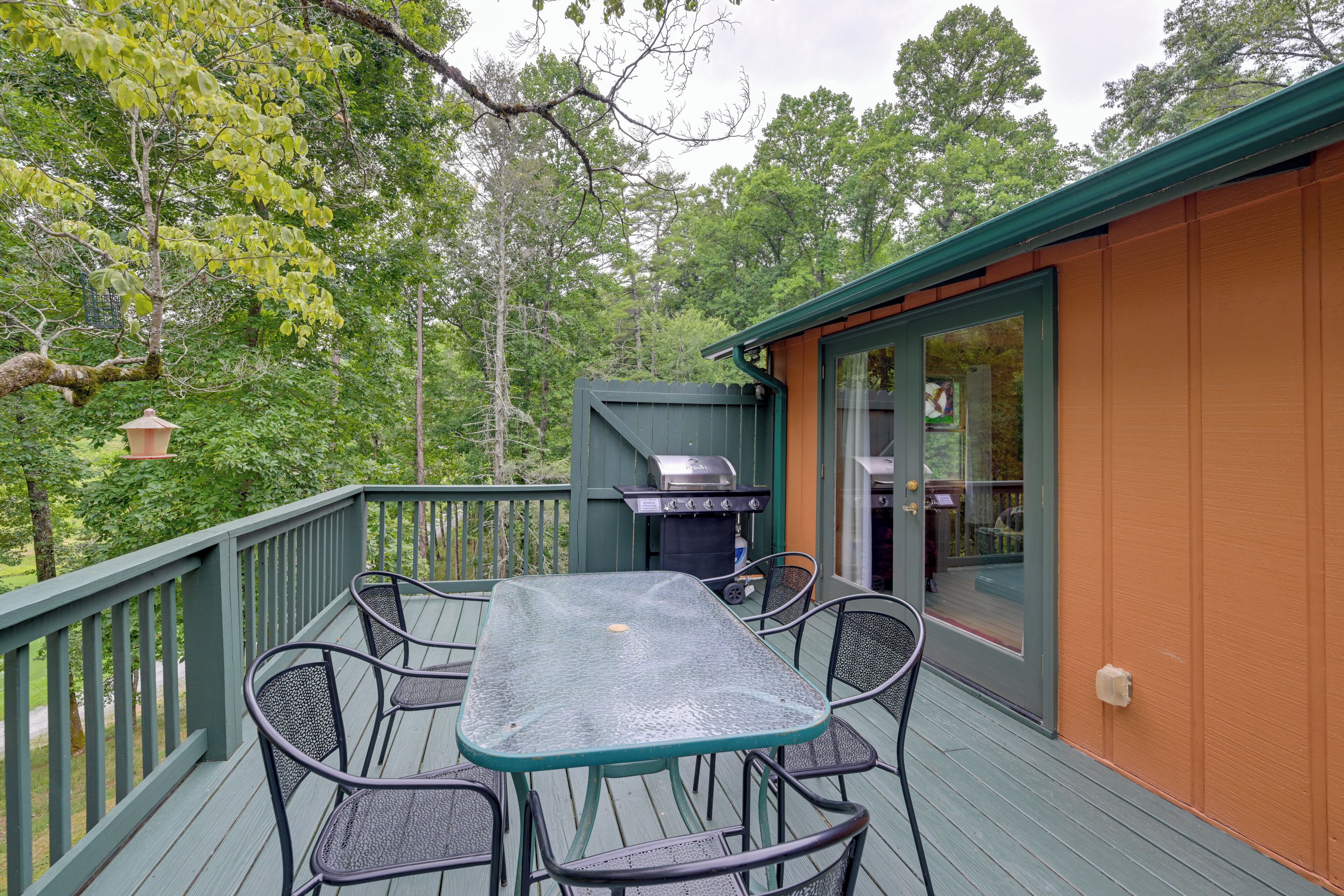 A covered outdoor deck with a table and chairs, a grill, and a view of the surrounding forest.