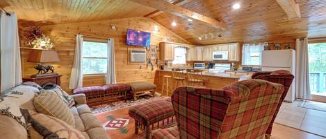 A cozy cabin interior with a wooden ceiling, a living room with a sofa and plaid chairs, a kitchen with wooden cabinets, and a television mounted on the wall.