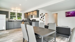 A modern kitchen with grey cabinets, a dining area with a table and chairs, and a TV mounted on the wall. - Mashpee Cottage w/ Deck: 6 Mi to South Cape Beach! (Mashpee)