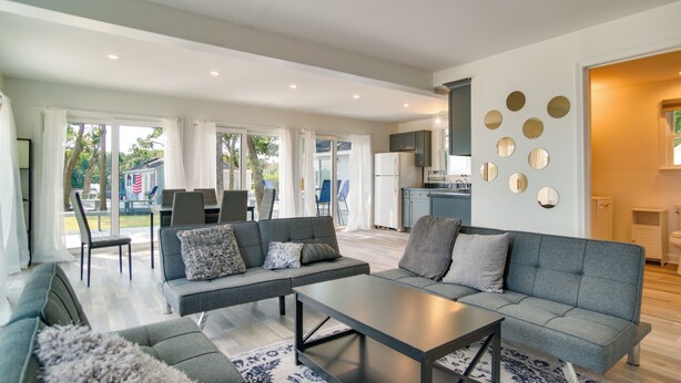 A modern living room with a grey sofa, a coffee table, and a patterned rug. There is a kitchen area with a refrigerator and cabinets. - Mashpee Cottage w/ Deck: 6 Mi to South Cape Beach! (Mashpee)