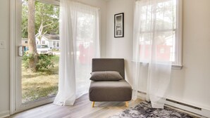 A room with a grey armchair, white curtains, a framed picture on the wall, and a view of a house and trees outside. - Mashpee Cottage w/ Deck: 6 Mi to South Cape Beach! (Mashpee)