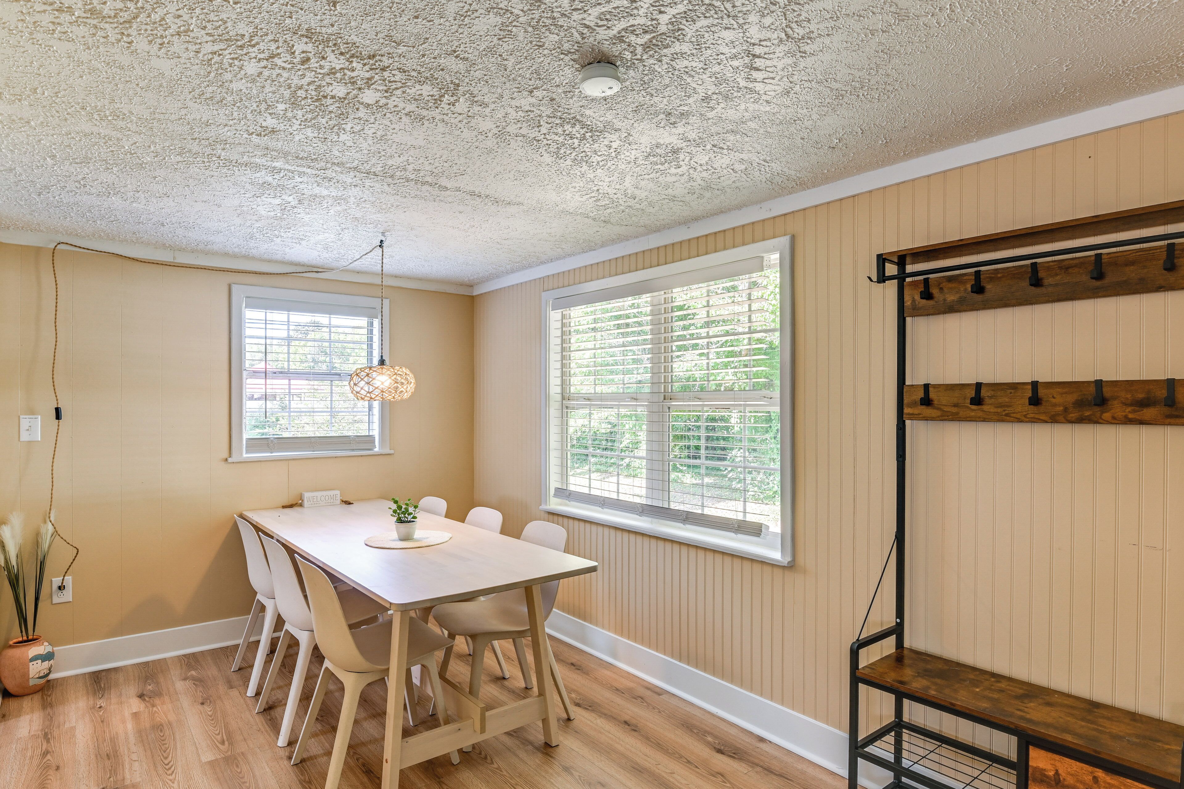 Un coin repas avec une table et des chaises en bois, deux fenêtres équipées de stores, un ventilateur de plafond et une étagère avec des crochets.
