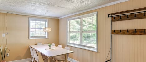 Un coin repas avec une table et des chaises en bois, deux fenêtres équipées de stores, un ventilateur de plafond et une étagère avec des crochets.