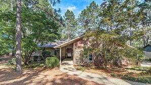 A single-story brick house with a front porch, surrounded by trees and a gravel driveway. - Charming Pinehurst Getaway w/ Golf Course View! (Pinehurst)