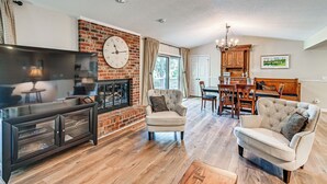 A living room with a fireplace, a television, and a dining area with a chandelier. - Charming Pinehurst Getaway w/ Golf Course View! (Pinehurst)