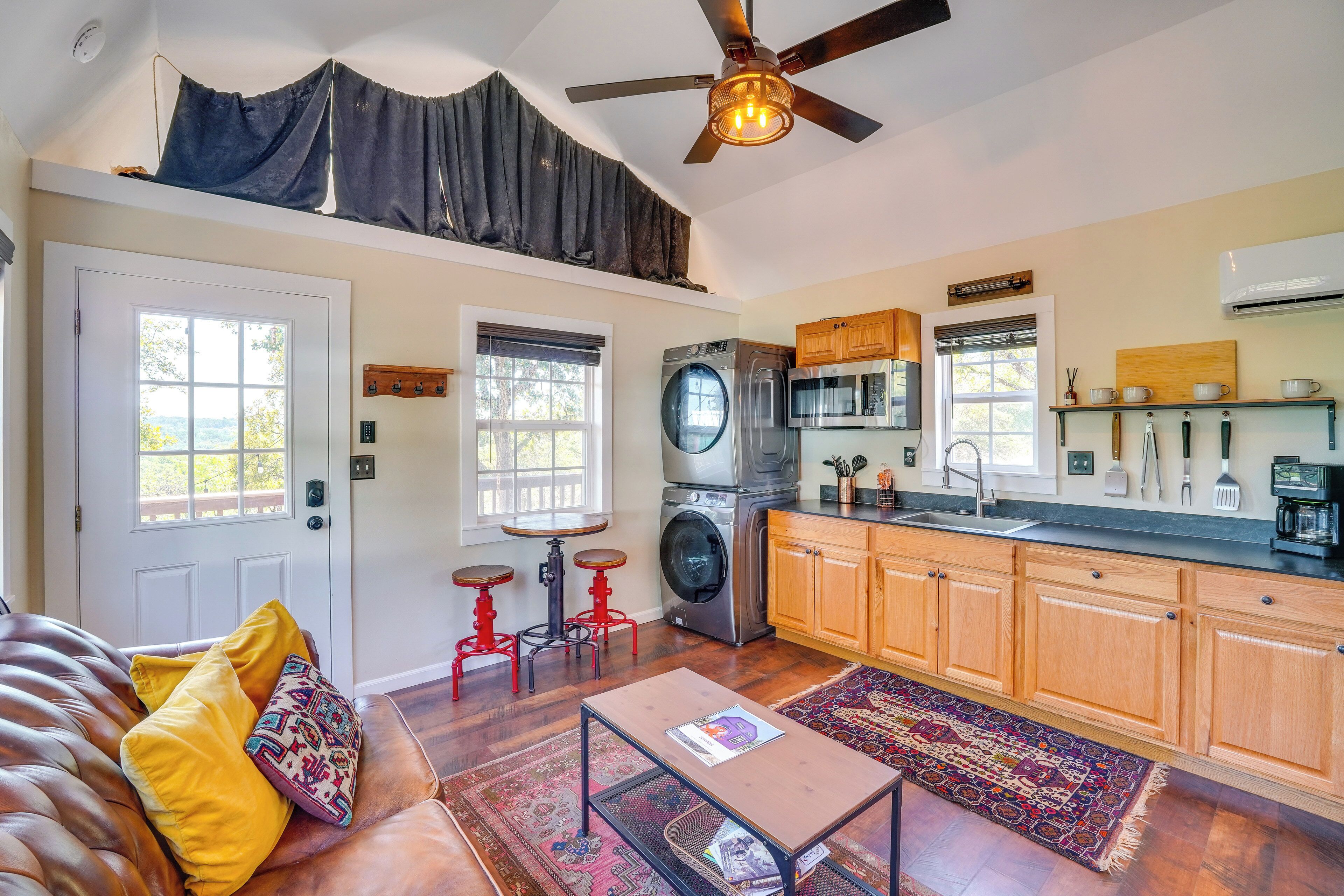 A cozy living room with a sofa, a coffee table, and a ceiling fan. Adjacent is a kitchen with wooden cabinets, a microwave, and a washing machine.