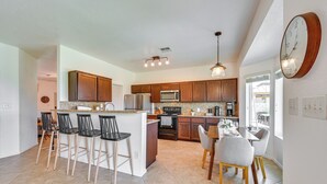 Uma cozinha com armários de madeira, uma ilha central com banquetas, uma mesa de jantar com cadeiras e um relógio na parede.
